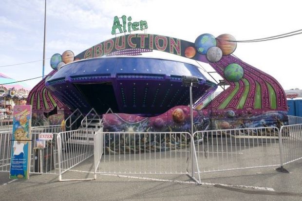 L’Alien Abduction est un manège de forme arrondie ayant une grande porte d’entrée; des lumières de couleur et des motifs du cosmos ornent le bas et la mascotte du WCA se trouve sur une affiche près du manège