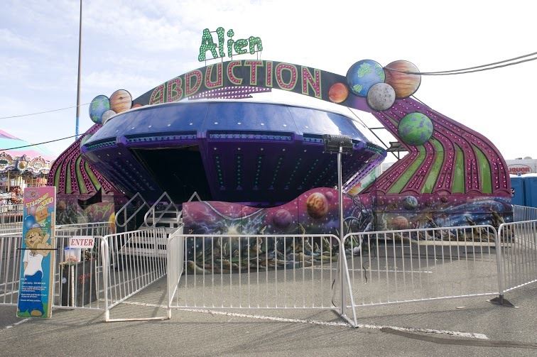 L’Alien Abduction est un manège de forme arrondie ayant une grande porte d’entrée; des lumières de couleur et des motifs du cosmos ornent le bas et la mascotte du WCA se trouve sur une affiche près du manège