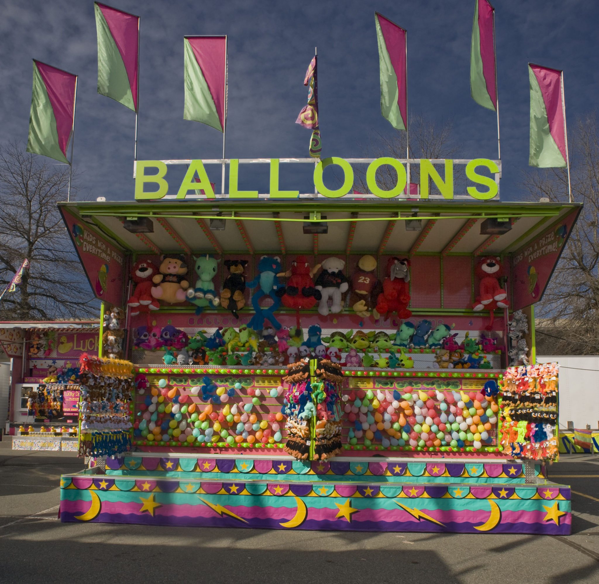 Des drapeaux flottent au-dessus d’un stand coloré de jeu de ballons à crever; des peluches sont suspendues au mur en haut des ballons et une toile aux couleurs pastels couvre le bas du stand