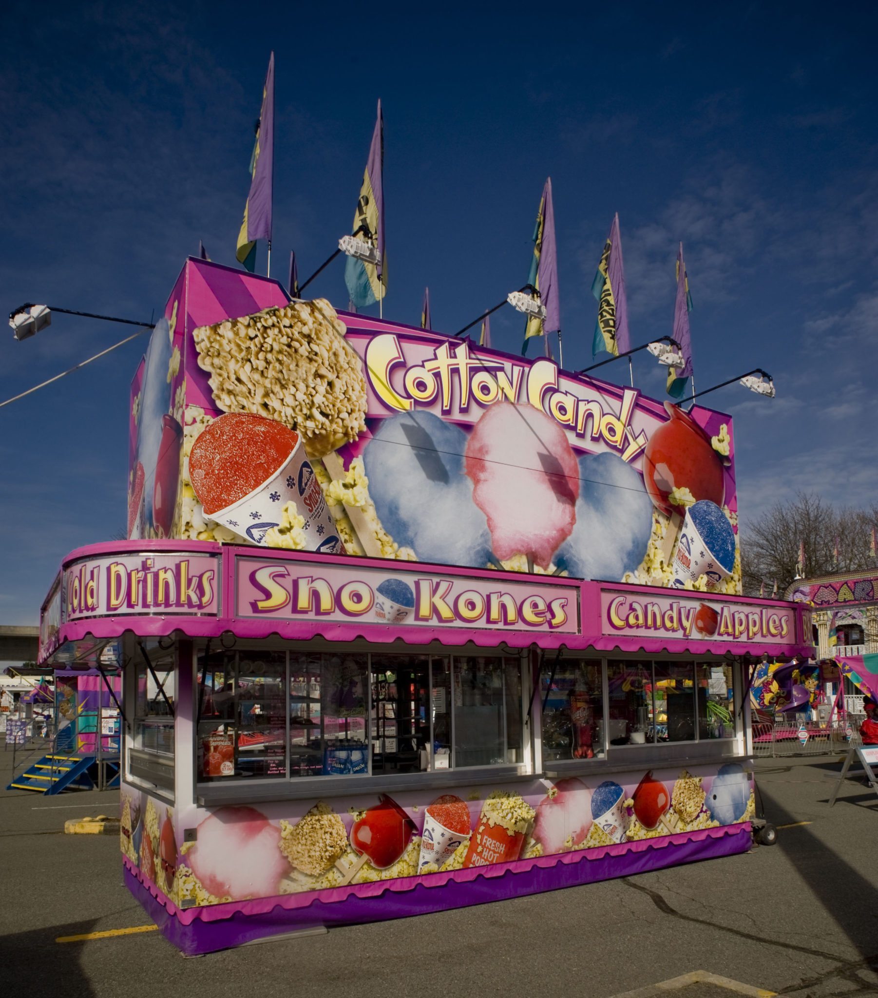 Un stand alimentaire offre de la barbe à papa, du maïs soufflé, des pommes glacées, des cornets de glace et des boissons froides; le stand est rose et violet, et est couvert de dessins colorés de gourmandises