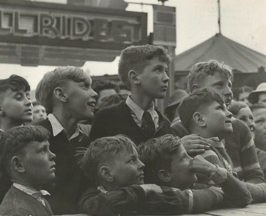 Une photo en noir et blanc en gros plan de jeunes garçons rassemblés à la fête foraine, en arrière- plan, on voit le panneau d’un manège à sensations fortes