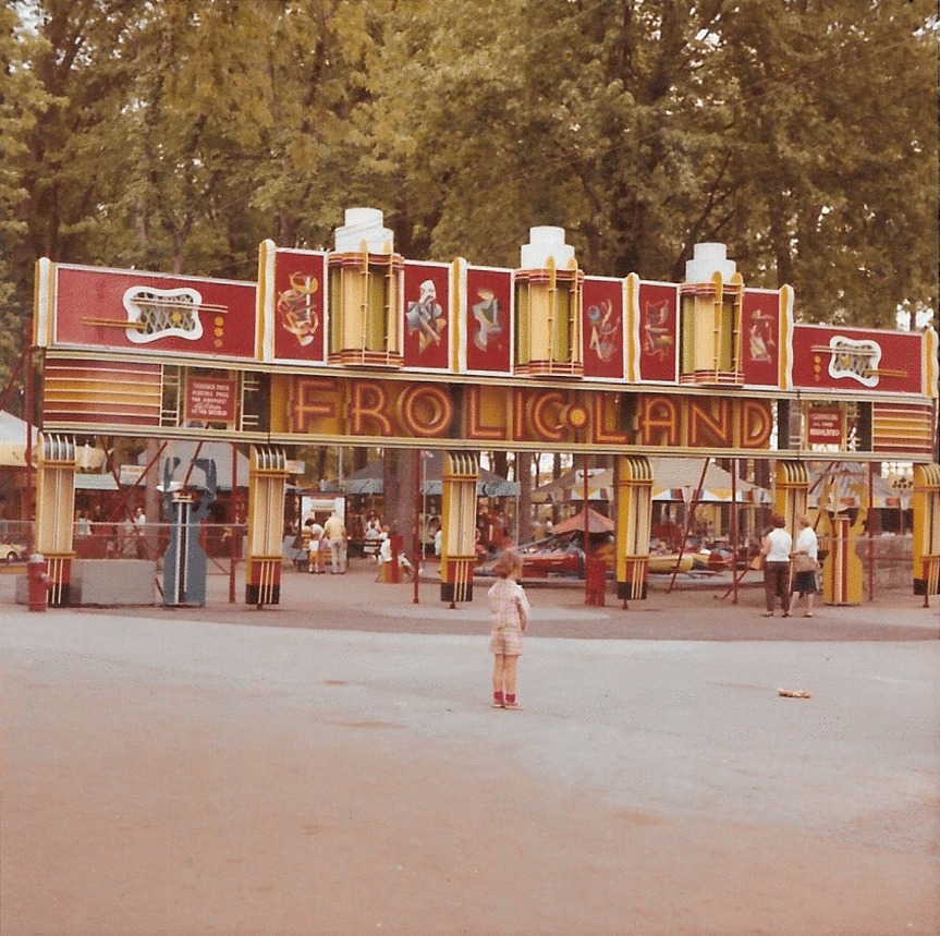 Une jeune fille est debout devant la grande entrée du Frolic Land, une attraction de la fête foraine; il n’y a personne d’autre autour