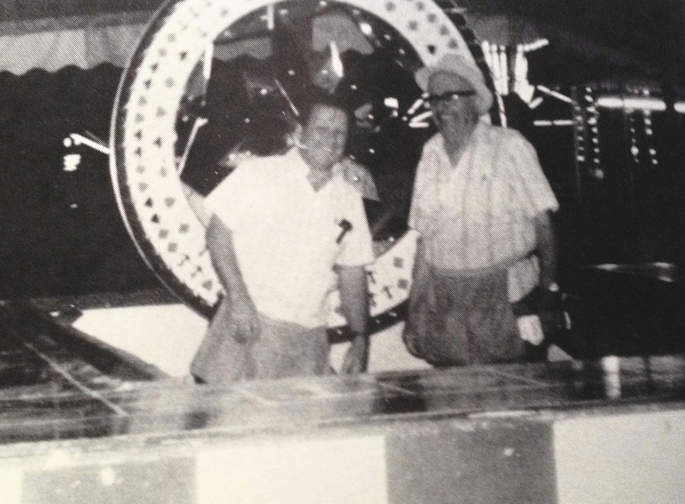 Photo en noir et blanc de Bingo Hauser, d’âge moyen, qui sourit et porte un casque-micro. Il est debout à côté d’un autre homme devant le comptoir d’un jeu de hasard et derrière eux se trouve une roue à faire tourner