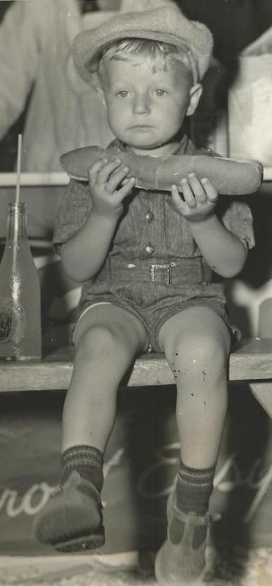 Une photo en noir et blanc d’un jeune enfant tenant un hot dog d’un pied de long, assis avec une bouteille de boisson gazeuse à côté de lui