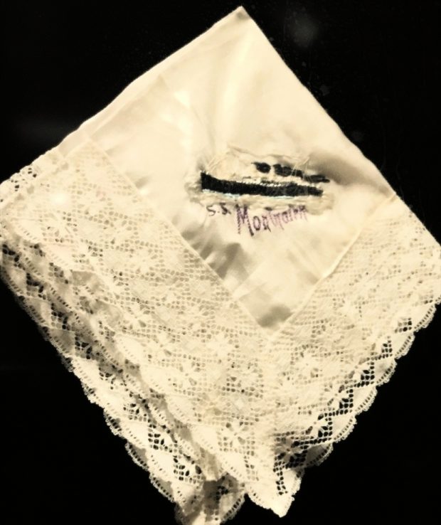 Un mouchoir blanc cassé plié et agrémenté d’une bordure en dentelle et d’une broderie noire avec des lettres violettes au centre