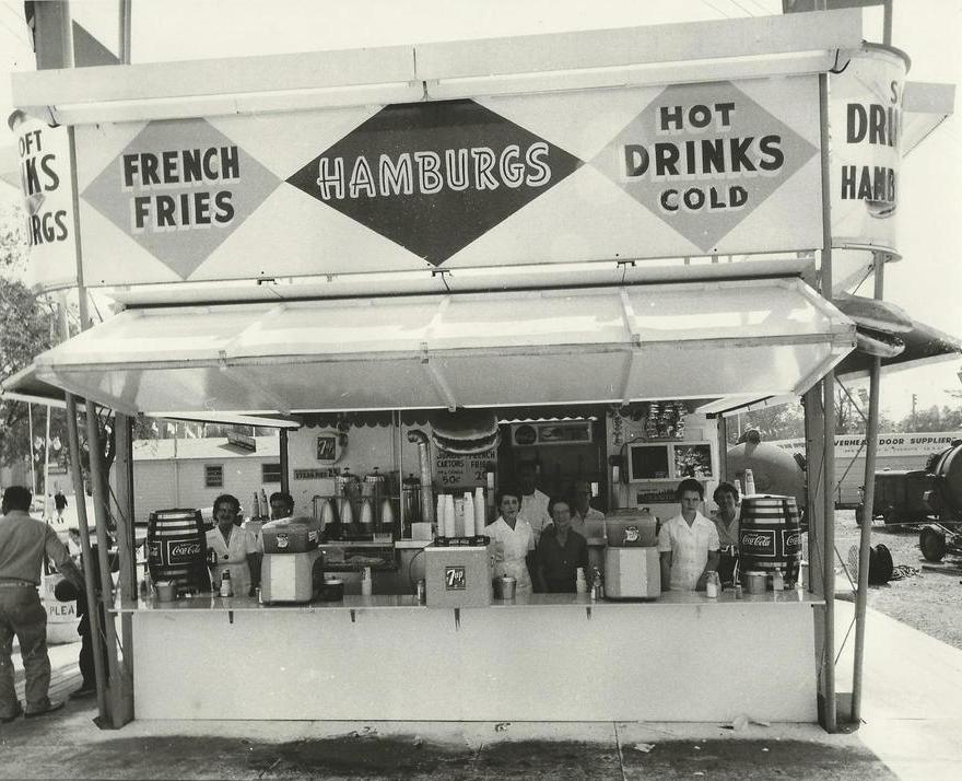 Une photo en noir et blanc d’un stand alimentaire, sur l’affiche il est écrit French Fries, Hamburgs and Cold Drinks (Frites, hamburgers et boissons froides); il y a des employés vêtus d’un uniforme tout en blanc à l’intérieur et à l’extérieur du stand