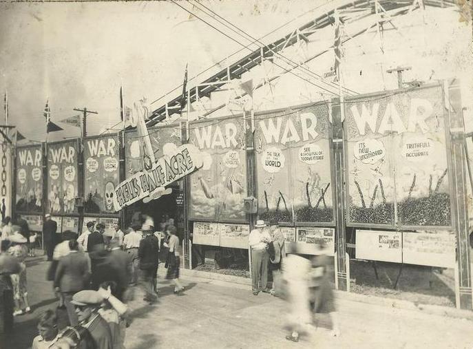 Une photo en noir et blanc d’une attraction devant laquelle se trouvent six panneaux sur lesquels on peut lire le mot War (guerre) et une affiche se balance au-dessus de l’entrée sur laquelle il est écrit « Hell’s Half Acre », un demi-acre d’enfer; des gens sont rassemblés devant l’entrée