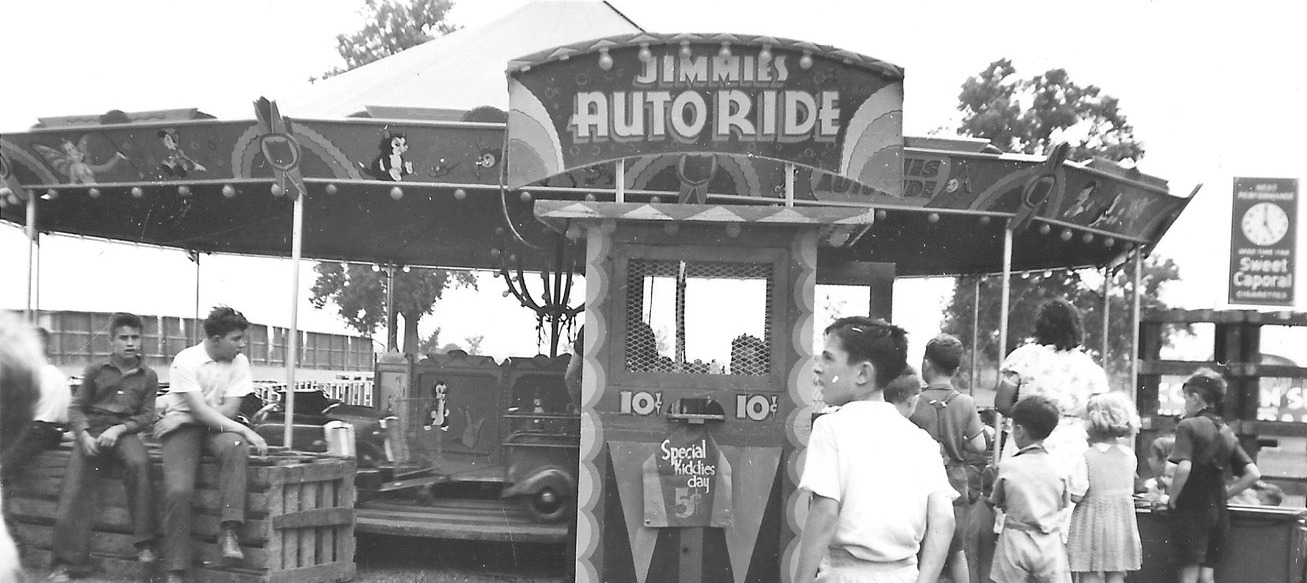 Une photo en noir et blanc de gens rassemblés autour d’un manège pour enfants; il y a un panneau sur lequel on peut lire Jimmie’s Auto Ride (manège les autos de Jimmie) et une affiche qui indique que c’est la journée des enfants à cinq cents (Special Kiddies Day five cents)