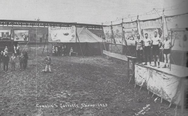 Une photo en noir et blanc de bannières de fête foraine et des hommes au torse nu sont debout sur une scène; il est écrit sur une bannière en arrière-plan les mots « physical culture », culture physique