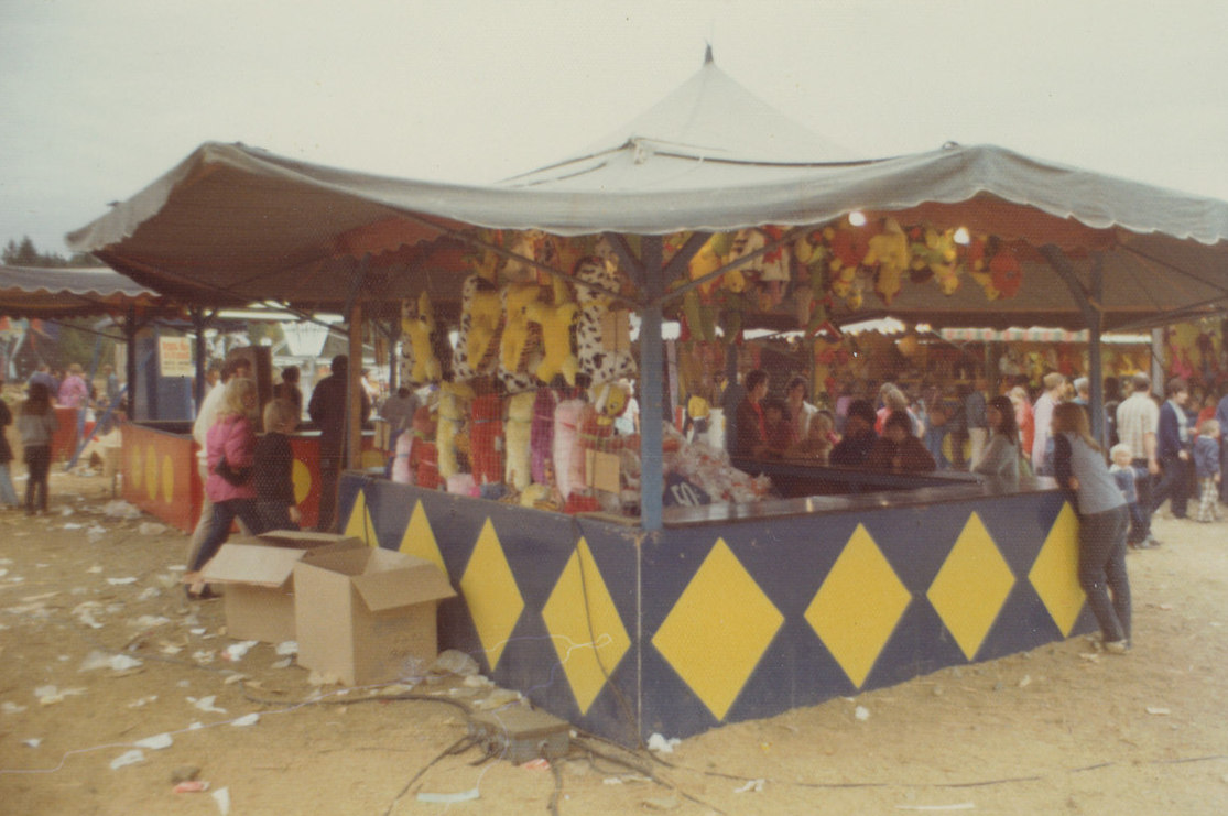 Dans une fête foraine, stand d’un jeu sous une toile, les prix, des animaux en peluche colorés sont suspendus tout autour du stand