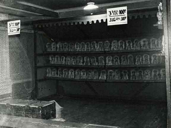 Une photo en noir et blanc d’un jeu d’adresse appelé « the cat rack », on voit un stand rectangulaire en métal avec trois rangées de clowns à faire tomber; il y a une affiche sur laquelle il est indiqué trois balles pour un dollar et trois chats pour trois dollars