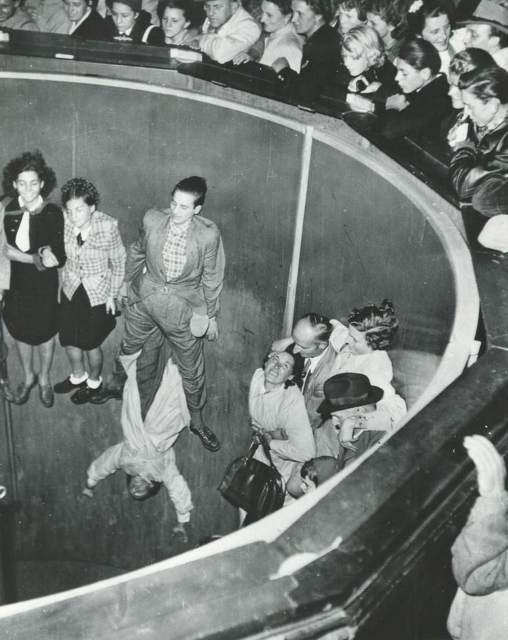Une photo en noir et blanc d’un grand manège à rotation, l’accélération glue les gens au mur d’une large cuvette ronde, plusieurs personnes situées en haut de la cuvette regardent les gens dans le manège