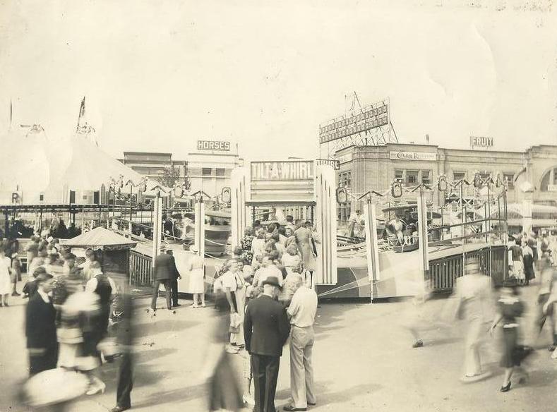 Une photo en noir et blanc d’une foule à une fête foraine et des gens à bord du manège « Tilt-a- Whirl »