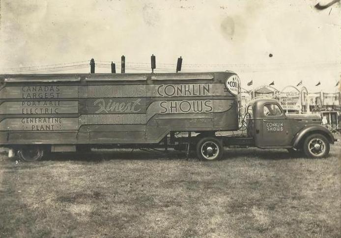Une photo en noir et blanc d’un grand camion de transport sur lequel il est écrit « Conklin Shows », et qui est muni d’un générateur portatif; en arrière-plan on voit des bannières de la fête foraine