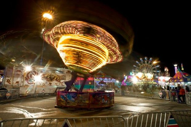 Dans une fête foraine, un manège illuminé tourne comme une toupie le soir et l’on voit une grande roue en arrière-plan