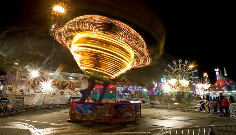 Dans une fête foraine, un manège illuminé tourne comme une toupie le soir et l’on voit une grande roue en arrière-plan