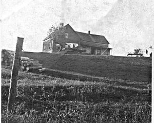 Photo d’archive en noir et blanc d’une maison de ferme située au sommet d’une colline, les bûches sont empilées à gauche jusqu’au milieu de la colline. Vers 1950.