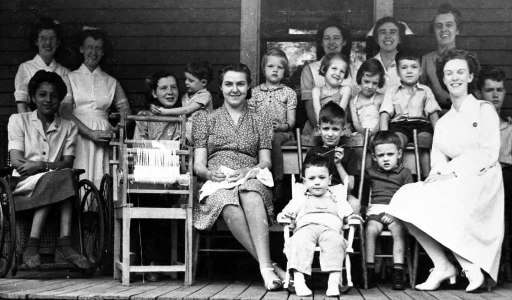 Photo de groupe en noir et blanc montrant des enfants, des infirmières et des membres du personnel assis sur une galerie; Edith Henry est assise dans un fauteuil roulant au premier rang, première à gauche.