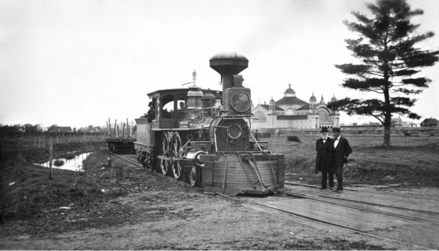 Photo d’archive en noir et blanc montrant une locomotive équipée du déblayeur d’entre-rails Temple & Miller, avec deux hommes portant un costume et un haut-de-forme qui se tiennent à droite.