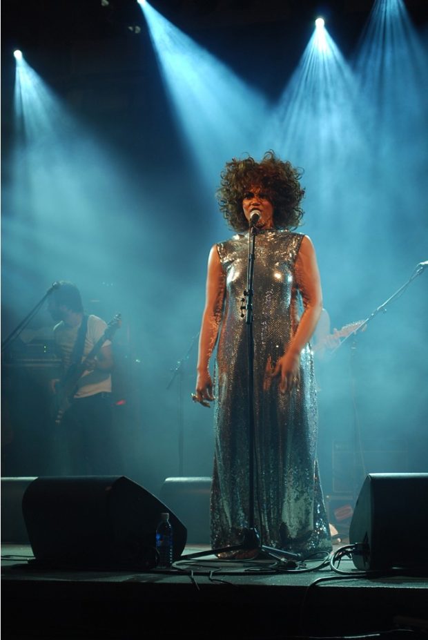 Photo récente d’une femme debout chantant sur une scène très éclairée et vêtue d’une robe longue. Un bassiste se trouve à l’arrière-plan.
