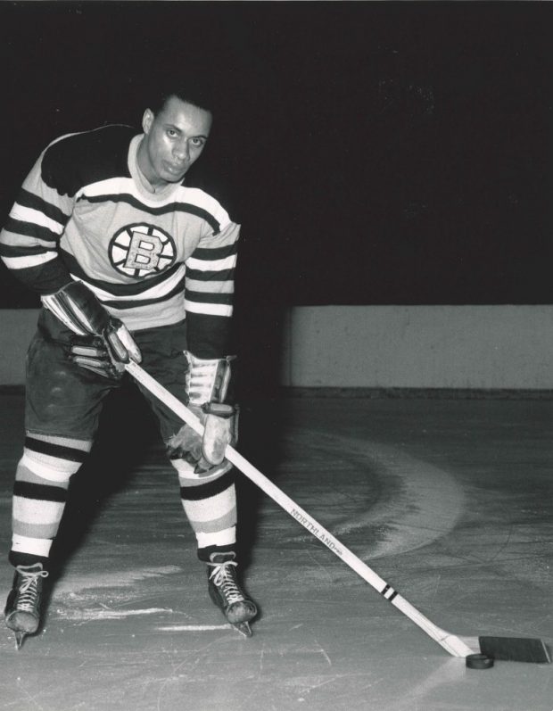 Photo en noir et blanc de Willie O’Ree sur la glace en tenue de hockey, portant l’uniforme des Bruins de Boston et tenant un bâton de hockey.