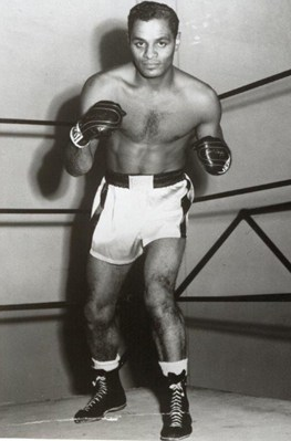 Photo en noir et blanc d’un homme noir dans une pose de boxe, vêtu d’un short et portant des gants de boxe.