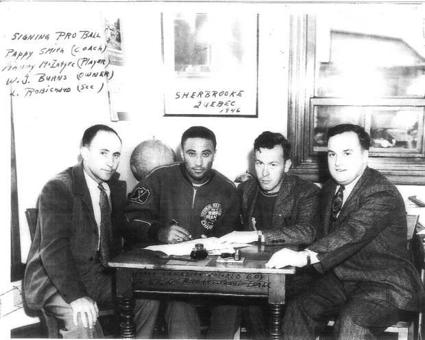 Photo en noir et blanc de quatre hommes assis à une table. Manny McIntyre, deuxième en partant de la gauche, signe un contrat de joueur de baseball professionnel.