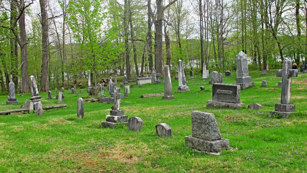 Photographie en couleurs d’un cimetière avec de l’herbe verte et plusieurs vieilles pierres tombales avec des arbres en arrière-plan.