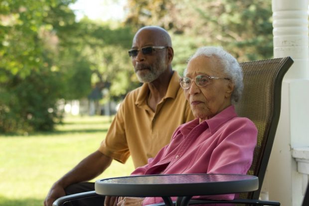 Photo en couleurs d’une femme noire âgée portant une chemise rose, assise. Derrière elle se trouve un homme noir portant une chemise jaune. 