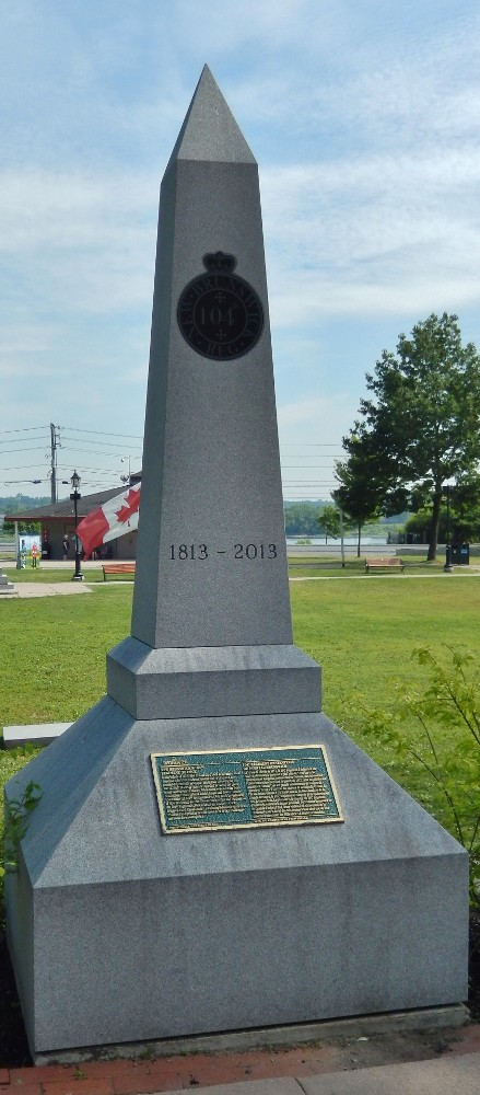 Photographie en couleurs du monument du 104e Régiment d’infanterie à l’entrée de la Place des officiers à Fredericton. À l’arrière-plan, on peut voir de l’herbe verte et des bâtiments du parc.