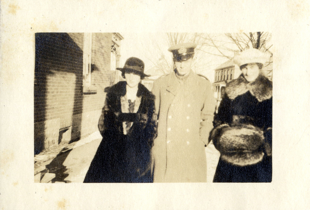 Deux femmes en manteau de fourrure et un homme en uniforme debout dans la neige devant une résidence.