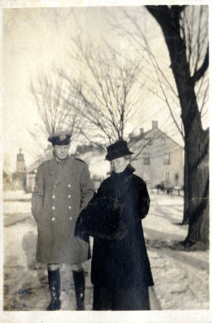 Un homme en uniforme et une femme en manteau de fourrure, debout dans la neige. On aperçoit des arbres et des maisons à l’arrière-plan.