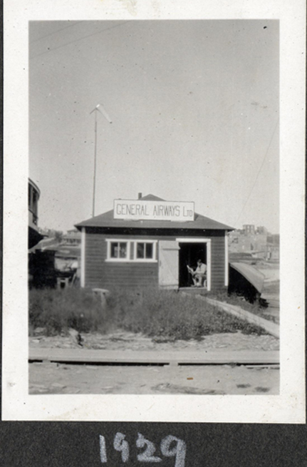 Petite bâtisse revêtue de bardeaux avec sur le toit une enseigne de la General Airways Ltd peinte à la main. À l’avant, un terrain mal entretenu et un trottoir en bois. Un homme lit, assis dans l’embrasure de la porte.