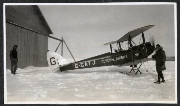 Deux pilotes en combinaison de vol devant un biplan monté sur skis, à côté d’un hangar.