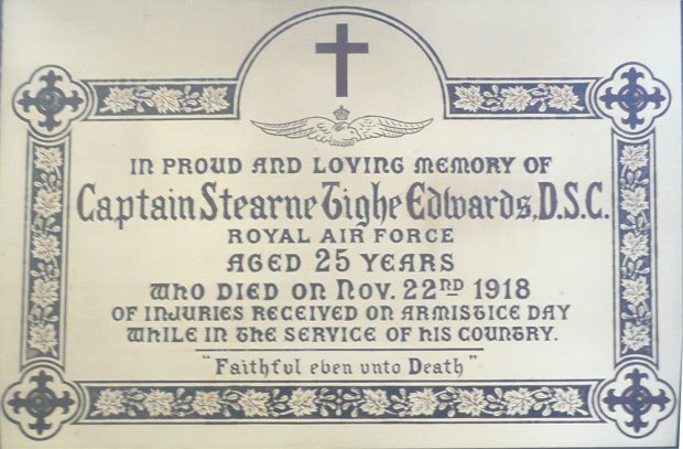 Plaque carrée, couleur cuivre, portant une inscription centrale bordée d’un motif de feuilles et de croix.