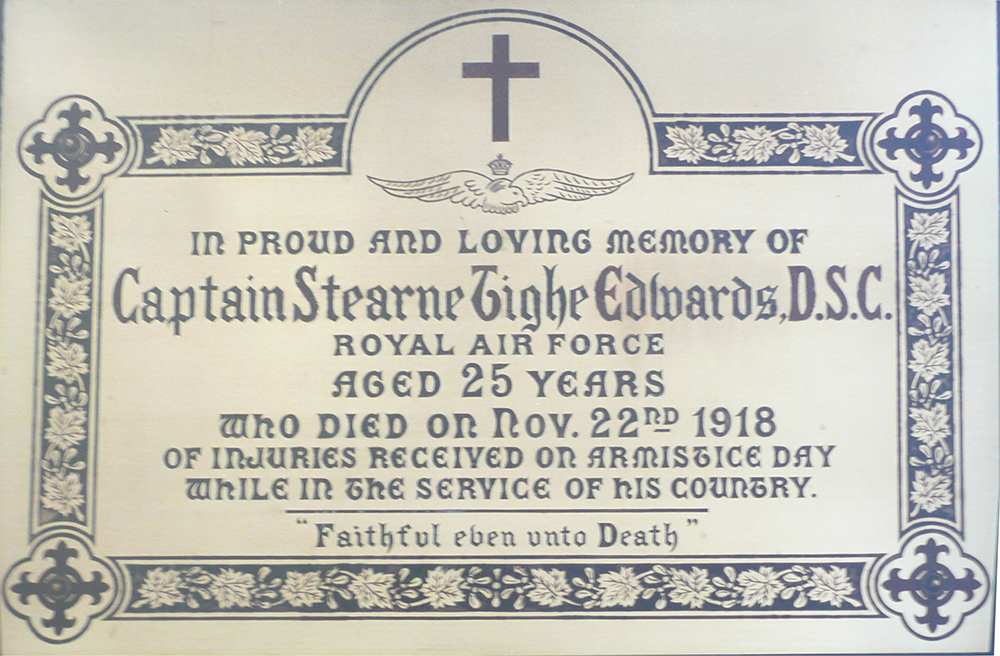Plaque carrée, couleur cuivre, portant une inscription centrale bordée d’un motif de feuilles et de croix.