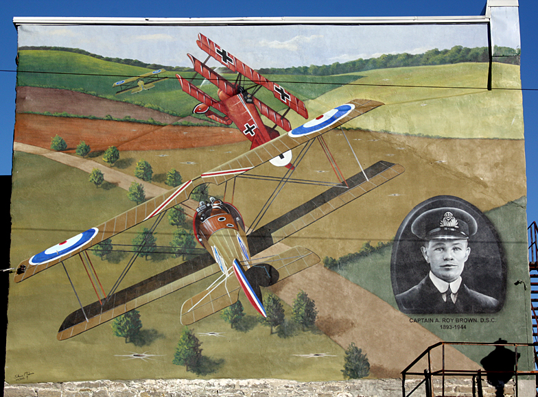 Deux biplans britanniques de la Grande Guerre et un triplan allemand combattent au-dessus des champs; portrait d’un homme dans le coin inférieur droit. 