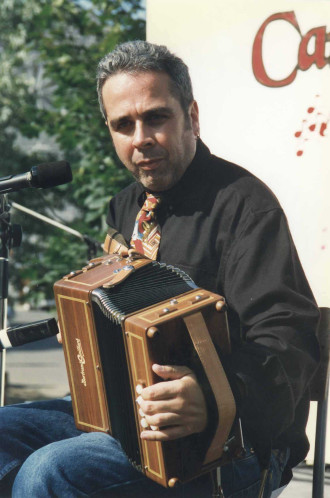 Photographie couleur, Alain Pennec, assis sur une chaise, son accordéon sur les genoux, regarde le photographe lors de la prise de la photo.