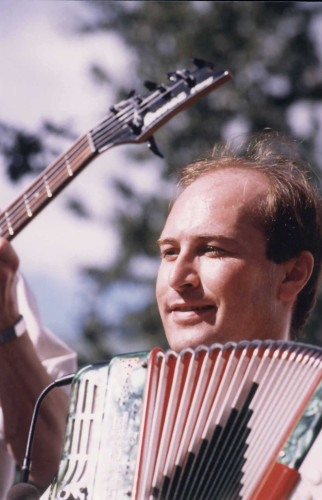 Photographie couleur, gros plan sur Bernard Félix et son accordéon. En arrière-plan, on aperçoit le manche d’une basse.