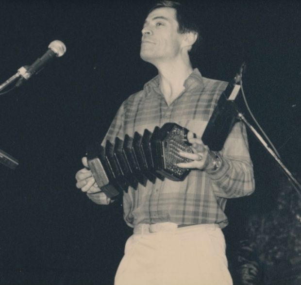 Photographie noir et blanc, Alistair Anderson son concertina en main, joue sur une scène intérieure. 