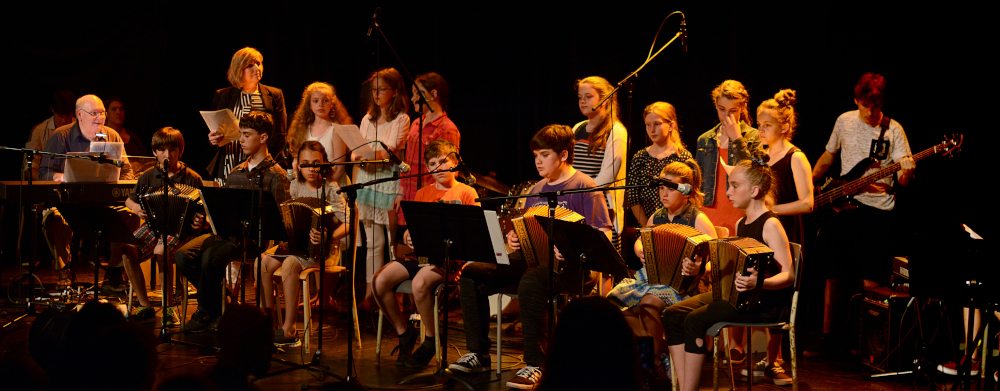 Photographie couleur, plusieurs jeunes musiciens d’âge scolaire livrent un concert sur une scène. 