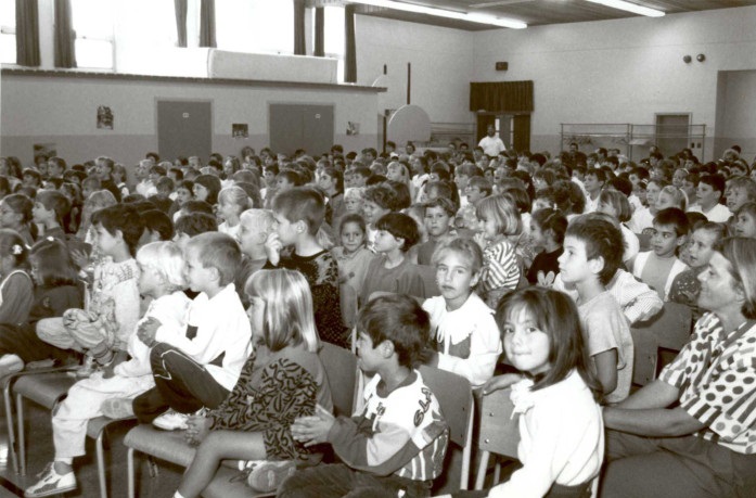 Photographie noir et blanc, assis sur des chaises, plusieurs groupes de jeunes enfants de l’école primaire sont réunis pour assister à un spectacle d’accordéon.