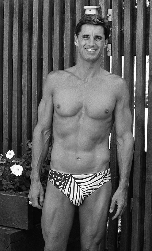  Portrait photo en noir et blanc du nageur Michael Mealiffe. Il porte des maillots de bain et se tient sur un patio extérieur avec une clôture à lattes en arrière-plan.