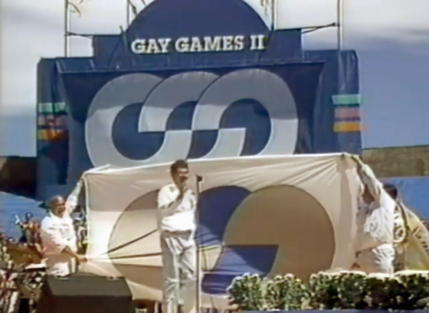  Lors des cérémonies de clôture des Gay Games II, Richard Dopson se tient sur la scène au micro devant un grand drapeau des Jeux gais tendu par la délégation de Vancouver.