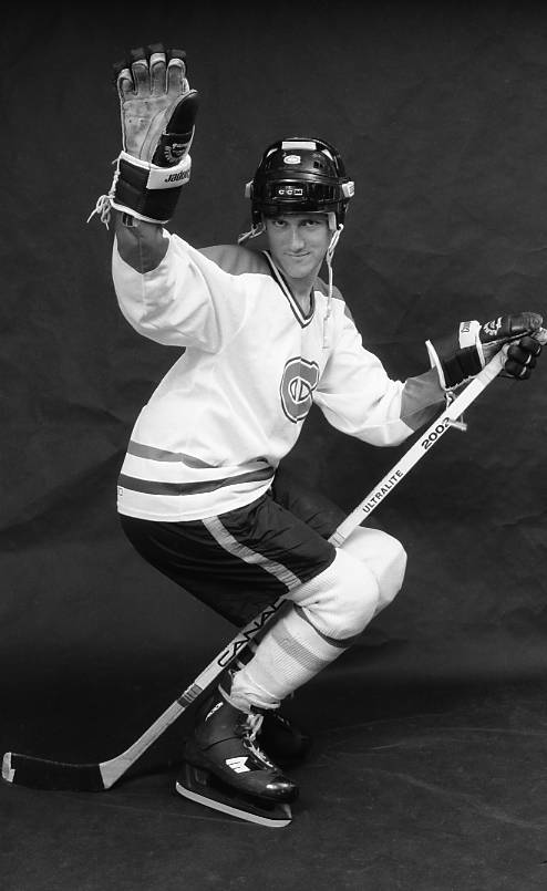 Photographie en studio en noir et blanc d'un joueur de hockey masculin portant un chandail blanc des Canadiens de Montréal et souriant en flirtant tout en se faisant passer pour «monter» son bâton.