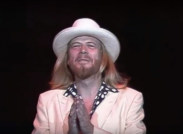  Le chanteur Long John Baldry, vêtu d'un costume blanc, d'une chemise noire à pois blancs et d'un fedora blanc, traverse la scène en direction du microphone en disant merci à la foule. Son groupe joue les premières mesures de A Thrill's A Thrill.