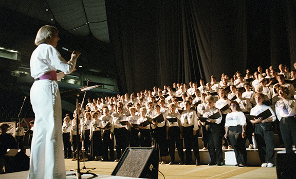  Plus de 70 membres du Celebration '90 Festival Chorus, en pantalon noir uniforme et chemises blanches, sont assemblés sur une colonne montante à plusieurs niveaux sur la scène de la Colombie-Britannique. Place Stadium. Carole White dirige leur prestation vocale lors des cérémonies de clôture.