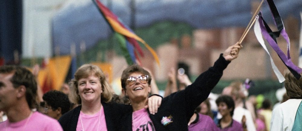  Un groupe d'athlètes féminines vêtues de t-shirts roses et de vestes noires sourit et salue la foule avec des banderoles colorées alors qu'elles marchent dans le défilé des athlètes lors des cérémonies d'ouverture. La gigantesque fresque de Vancouver qui soutenait la scène est visible en arrière-plan.