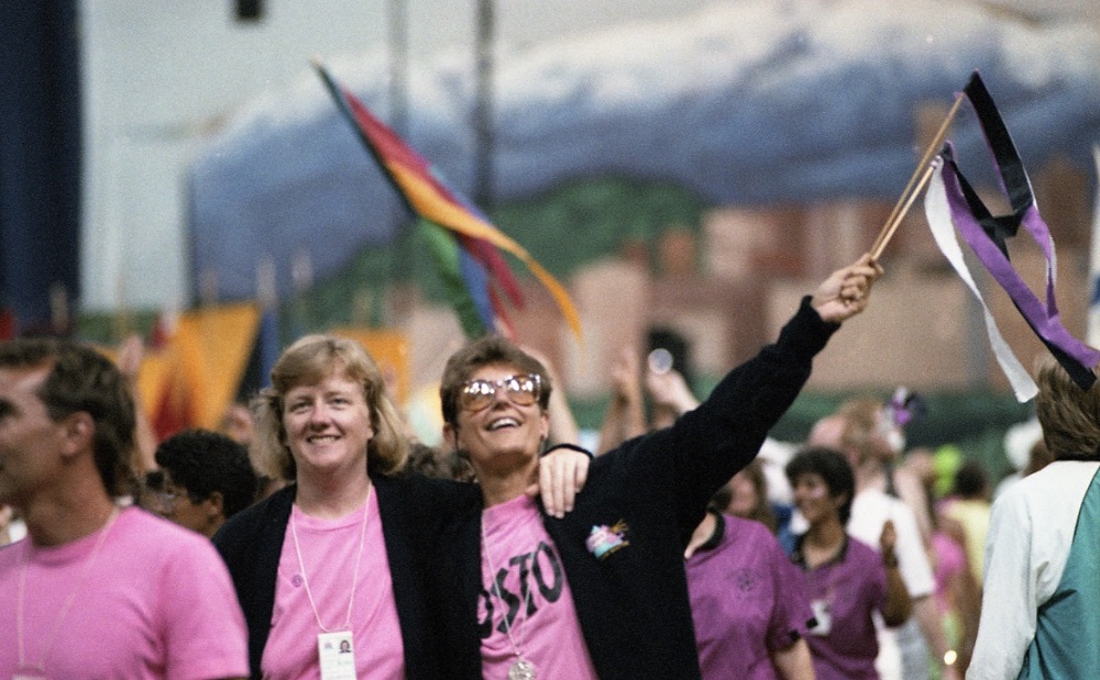  Un groupe d'athlètes féminines vêtues de t-shirts roses et de vestes noires sourit et salue la foule avec des banderoles colorées alors qu'elles marchent dans le défilé des athlètes lors des cérémonies d'ouverture. La gigantesque fresque de Vancouver qui soutenait la scène est visible en arrière-plan.