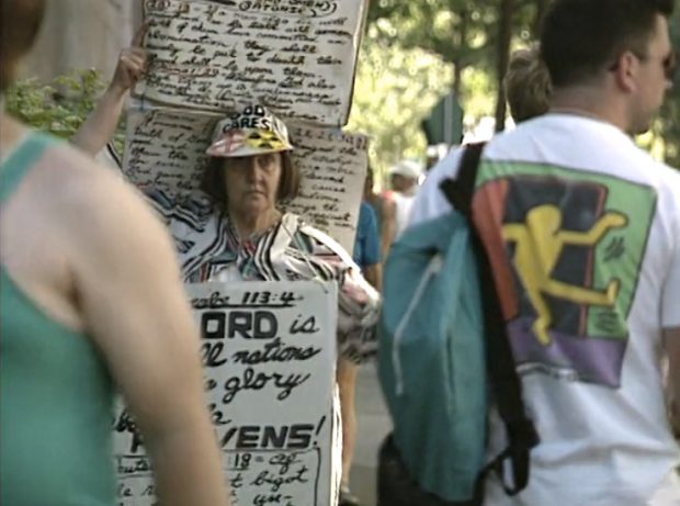 Une femme portant plusieurs panneaux d'affichage avec des passages bibliques écrits à la main et des discours homophobes sur eux se déplace à travers une foule d'inscrits aux Jeux gais.